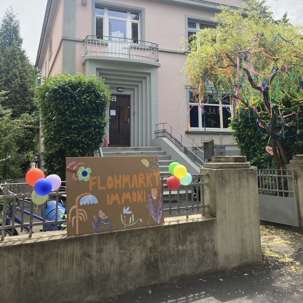 Die Villa des Kinderhauses mit einem bunt bemalten Schild „Flohmarkt im Moki“ und Luftballons am Zaun