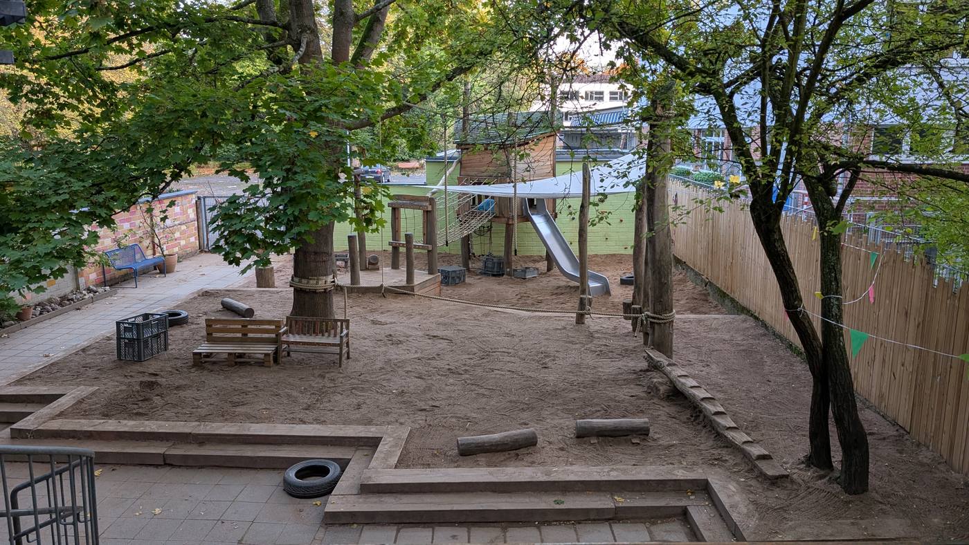 Blick über den Garten mit Sandfläche, großen Bäumen, Spielhaus, Kletternetz und Rutsche